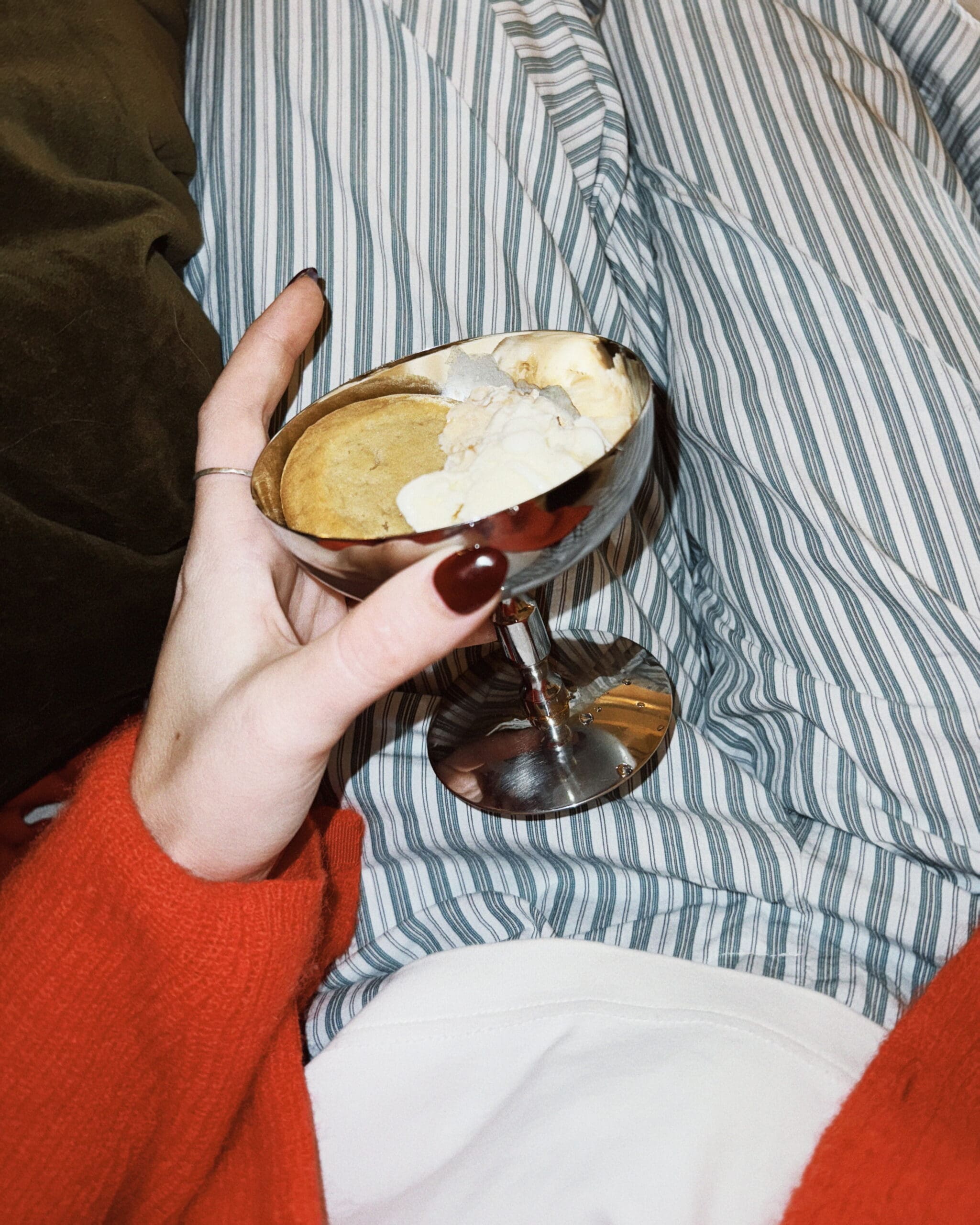 A cozy hand holding a silver coupe glass filled with a warm cookie and melting ice cream, resting on striped bedding. A red sweater sleeve adds warmth to the relaxed scene.