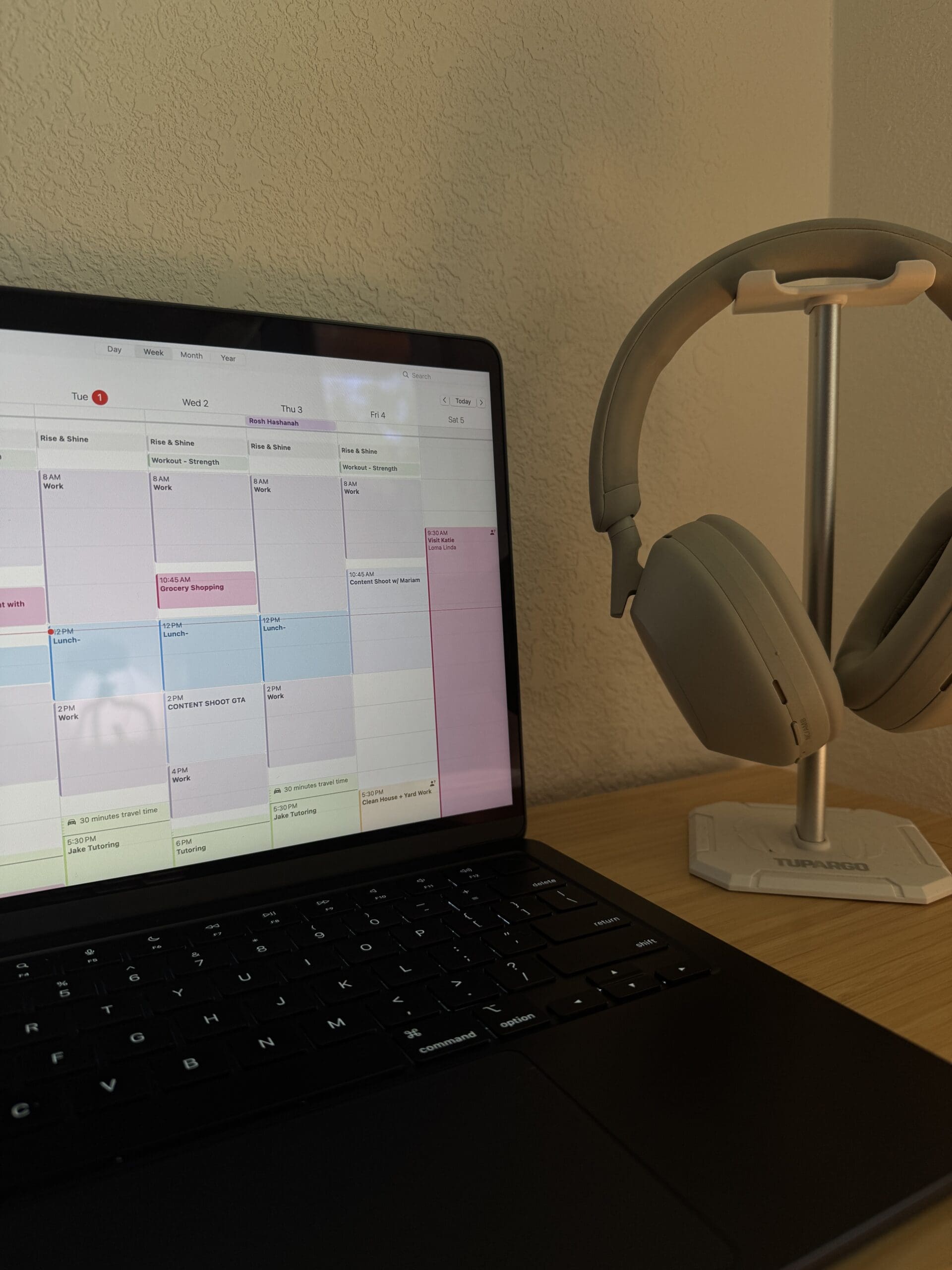 A MacBook displaying a time-blocked digital calendar sits on a wooden desk next to a pair of beige over-ear headphones on a stand, representing a well-organized and productive workspace.